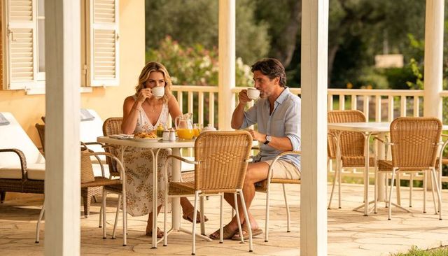 Couple sipping coffee on sunlit veranda, woman in floral sundress, man in linen shirt
