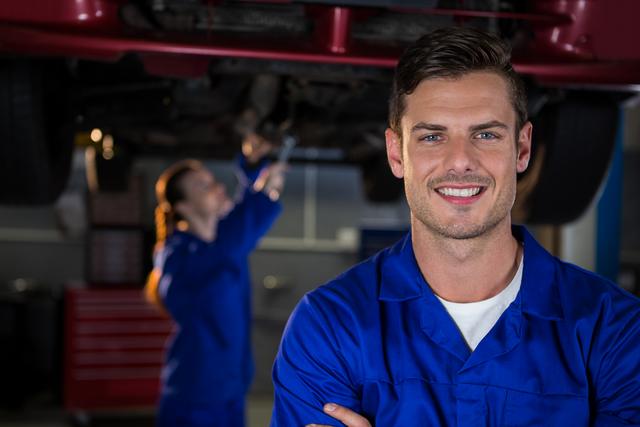 A happy mechanic in a blue uniform standing with arms crossed in a repair garage. In the background, another mechanic is working on a car. This image can be used for promoting automotive services, repair shops, and professional mechanic services. It conveys confidence, professionalism, and expertise in car maintenance and repair.