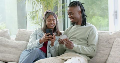 Diverse couple making online payment with smartphone and credit card on cozy sofa