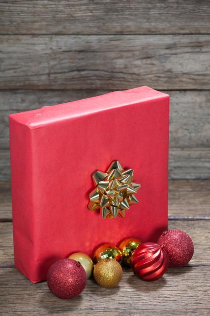 Red gift box with gold bow and assorted baubles on wooden table. Ideal for holiday promotions, festive greeting cards, Christmas advertisements, and seasonal blog posts.