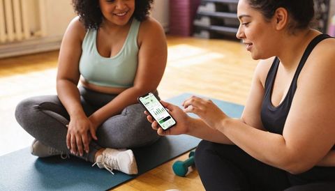 Two women sharing fitness app progress on smartphone while resting on yoga mats