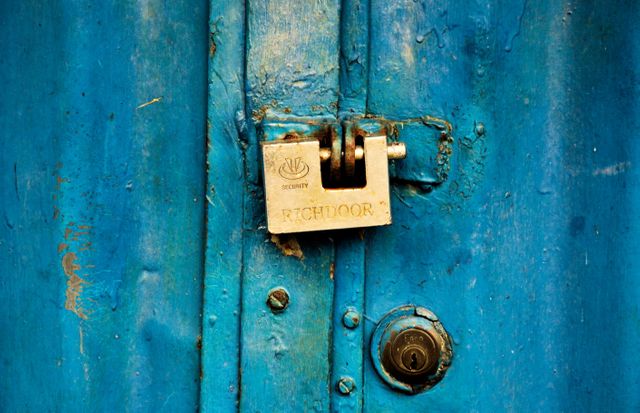 Close-up of a rusty padlock securing a weathered blue metal door. Perfect for themes related to security, antiquity, and rustic charm. Useful for websites, blogs, and publications covering topics on home security, vintage decor, and abandoned places.