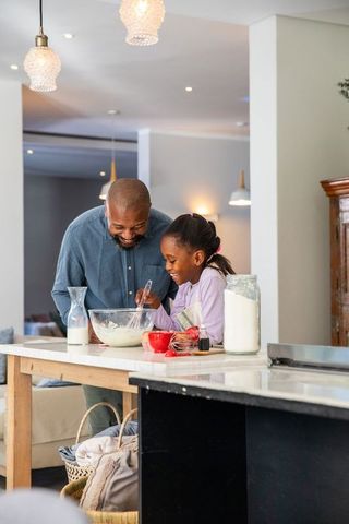 African american father and daughter whisking batter at kitchen island, joyful home baking