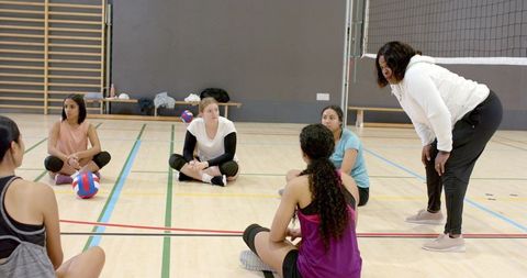 Female volleyball team sitting in semicircle listening to coach during indoor training