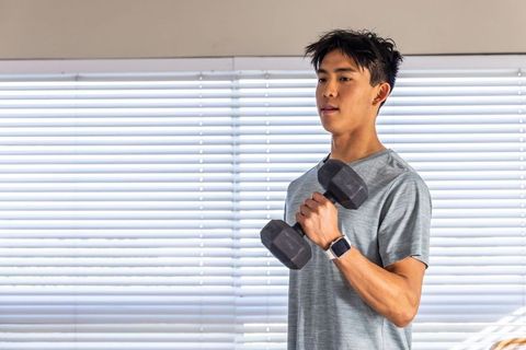 Asian man lifting hex dumbbell wearing smartwatch during home workout by window blinds