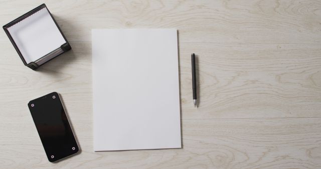 Blank paper lying on a wooden desk with a pen, note holder, and smartphone nearby. Suitable for workspace backgrounds, office setup designs, highlighting minimalistic and organized work environments, creating content for productivity and organizational blogs.