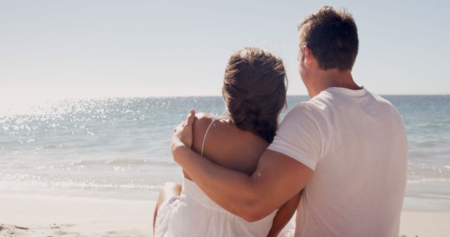 Couple sitting closely on sandy beach with tranquil sea background. Both are wearing casual attire and sharing a comforting embrace while gazing at the water. Ideal for concepts related to love, romance, vacations, relaxed lifestyles, and intimate moments on scenic beaches.