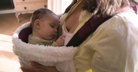Mother cradling sleeping baby in cozy carrier indoors