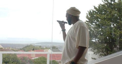 African man on balcony wearing beanie speaking into smartphone with coastal suburban view