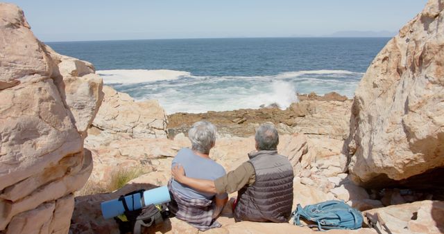 Back view of diverse senior couple sitting on rocks and looking at sea. Retirement, vacations, togetherness, summer, active senior lifestyle, unaltered.
