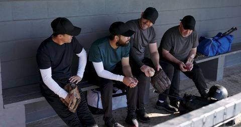 Diverse baseball team sitting on dugout bench holding gloves and bat sharing camaraderie