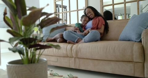 African american mother cuddling child on sofa, using smartphone, cozy living room
