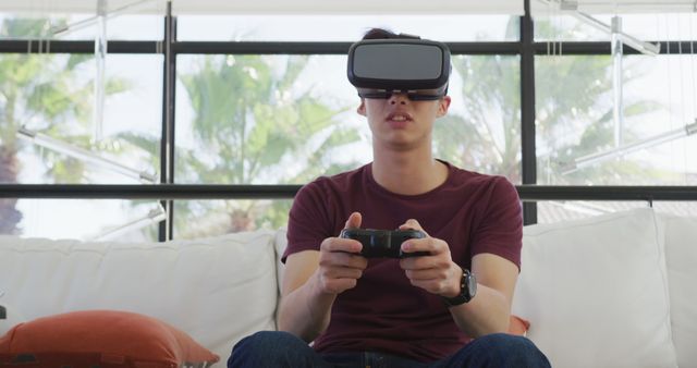 Young man wearing VR headset while gaming on a couch, illustrating the immersive experience of virtual reality in home entertainment. Ideal for topics on modern technology, gaming culture, and VR innovations. Suitable for articles, blogs, and advertisements related to consumer electronics, gaming, and tech lifestyle.