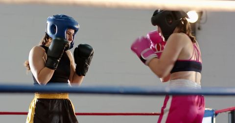 Two Female Boxers Sparring in Ring Wearing Headguards and Gloves Under Gym Lighting