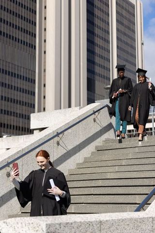 Three graduates taking selfie on city steps with diplomas and mortarboards, urban celebration