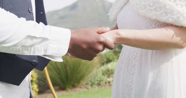 Image of midsection of diverse bride and groom holding hands and walking at outdoor wedding. Marriage, love, happiness and inclusivity concept.