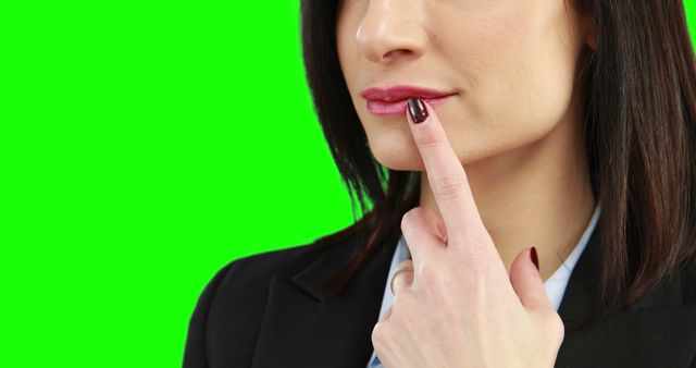 A Caucasian businesswoman is touching her lips thoughtfully, with copy space on a green chroma key background. Her contemplative gesture suggests she is pondering a decision or idea.