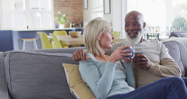 A senior couple is sitting on a sofa, enjoying hot drinks in a cozy living room. Both appear content and engaged in conversation. This can be used for themes such as retired life, togetherness, home comfort, elderly lifestyle, and quality time.