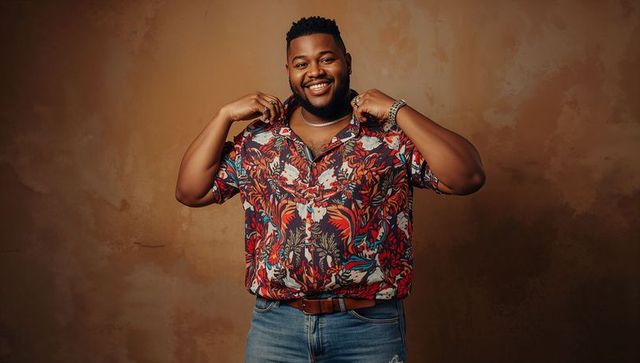 Smiling man adjusting floral shirt, wearing chain and watch, posing against brown textured backdrop