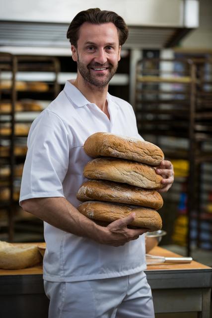 A professional baker is smiling while holding a stack of freshly baked bread loaves in a bakery. The background shows a busy kitchen with racks of bread. This image is ideal for use in articles or advertisements related to baking, culinary arts, professional chefs, bakery businesses, and food industry promotions.