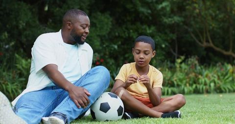 African american father coaching son with soccer ball on garden lawn, outdoor bonding