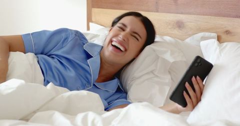 Woman Wearing Blue Pajamas Laughing While Using Smartphone in Bed