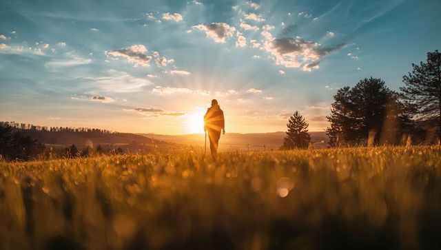 Silhouetted hiker walking into golden sunrise across dew-covered meadow with sunbeams