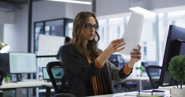 Young businesswoman wearing glasses, deeply focused on reviewing documents in a modern office environment. Ideal for corporate, professional, and business-related content to depict analysis, concentration, and workplace settings.