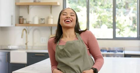 Laughing woman leaning on marble kitchen island wearing olive apron and smartwatch