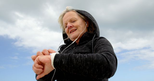 Senior woman in a black hoodie and earphones checking her smartwatch while exercising outdoors under a cloudy sky. This image is perfect for promoting healthy lifestyles, fitness technology, and outdoor activities for elderly.