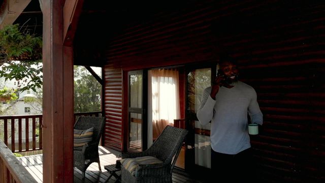 Man enjoying tranquil time on log cabin veranda, sipping tea and using a smartphone. Warm natural setting suggests relaxation and a rural lifestyle, with opportunities for connecting digitally. Ideal for themes related to slow living, woodland holidays, and digital communication in nature.