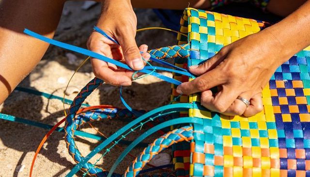 Closeup hands braiding blue straps on colorful woven beach tote under warm sunlight