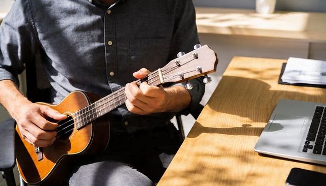 Man playing ukulele fretting chords at sunlit desk with laptop and papers