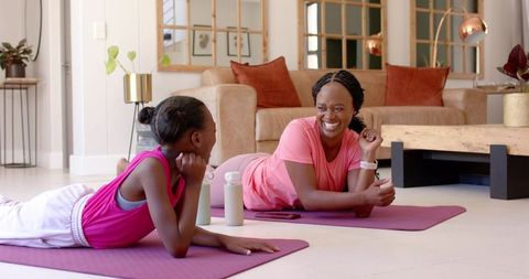 African american mom and teen daughter working out, smiling while holding plank on yoga mats