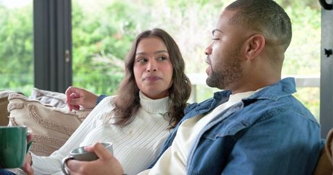 Diverse couple relaxing on sofa drinking coffee and sharing quiet morning conversation