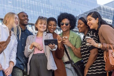 Diverse professional team laughing and sharing smartphone outside modern glass office plaza
