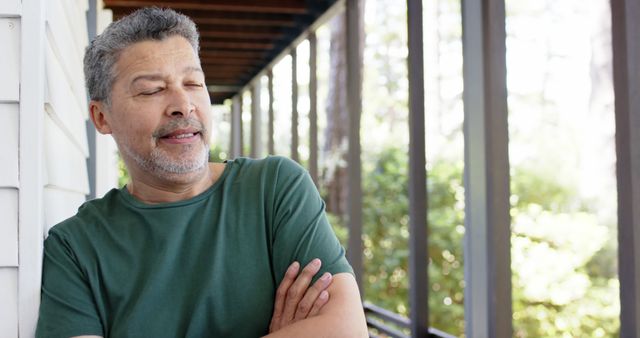 Middle-aged man leaning against a wall on a porch with a relaxed expression and arms crossed. Great for uses depicting leisure, relaxation, home life, and mature contentment.
