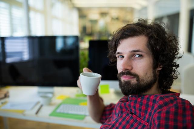 Young male graphic designer with curly hair and beard drinking coffee at his desk in a modern office. He is wearing a red plaid shirt and sitting in front of a computer. This image can be used for promoting creative work environments, office culture, or coffee breaks at work.