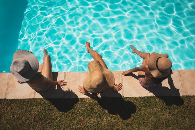 Three women are sitting by the edge of a swimming pool, wearing large hats and enjoying the summer sun. The clear blue water and bright sunlight create a refreshing and relaxing atmosphere. This image is perfect for promoting summer vacations, outdoor activities, leisure time, and friendship. It can be used in travel brochures, lifestyle blogs, and advertisements for summer products.
