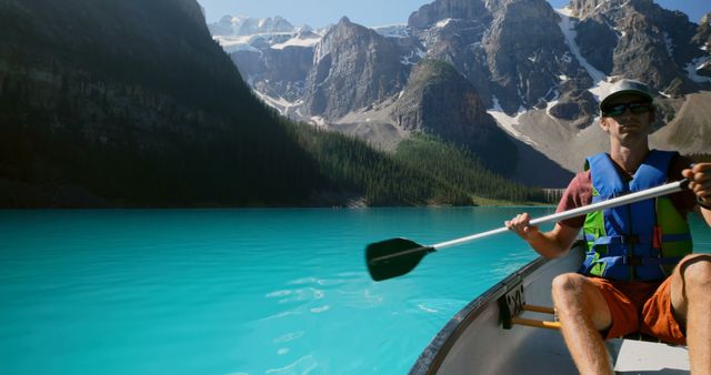 Man canoeing on a turquoise-colored lake surrounded by a dramatic mountain range, under clear blue skies. Ideal for use in adventure travel promotions, outdoor activity advertisements, nature exploration themes, and vacation destination marketing materials.