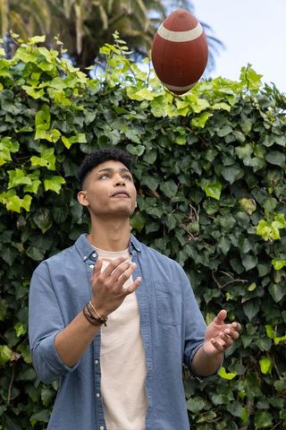 Young man catching football in backyard garden wearing chambray shirt and bracelets