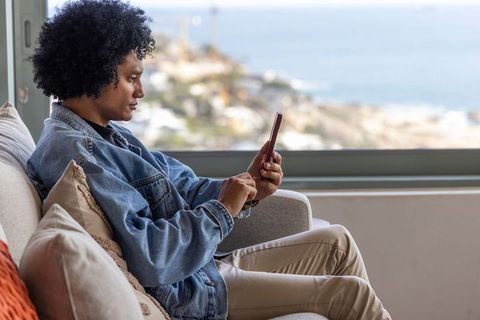 Relaxing african american man using smartphone on sofa with coastal window ocean view
