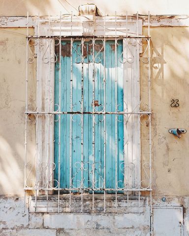 Weathered turquoise shutter behind ornate white iron grille on sunlit rustic wall