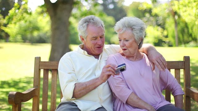 Elderly couple smiling while sitting on bench in scenic park, using digital camera. Can be used in content focusing on active retirement, relationships, or enjoying outdoor activities. Ideal for promotions highlighting senior-friendly technology or leisure activities for older adults.