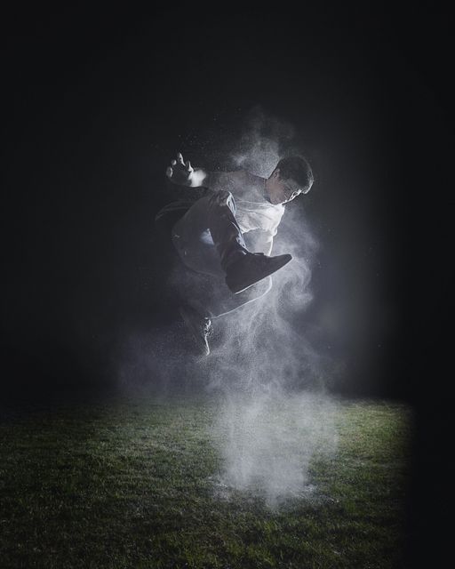 Young man performing an explosive mid-air jump at night with dust particles around, creating a dynamic and energetic effect. Scene set on grass with a dark sky backdrop. Useful for fitness, dance, adrenaline sports promotions, and dynamic movement themes.