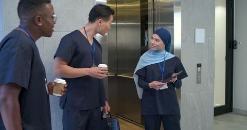 Diverse medical team standing and collaborating by elevator holding tablet and coffee