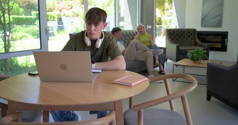 African american teen working on laptop at round table while family relaxes in living room