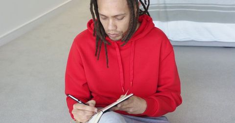 African american man dreadlocks sitting writing in notebook wearing red hoodie at home