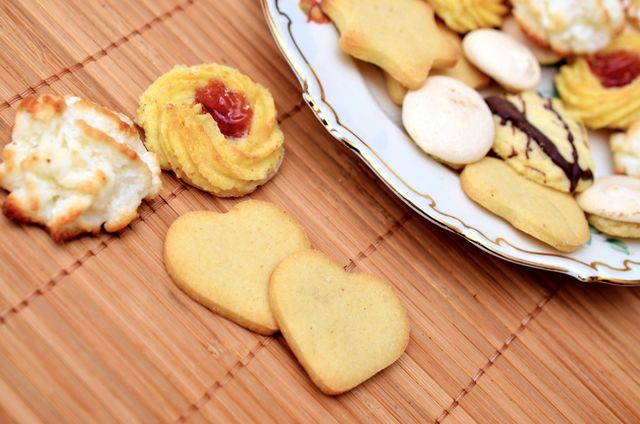 Assorted cookies and pastries displayed on a decorative plate placed on a wooden mat. Different types of cookies, including heart-shaped ones, are arranged making it ideal for showcasing a variety of baked goods, culinary presentation, and promoting dessert recipes or bakery products.