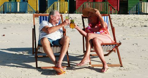 Senior Couple Toasting Drinks in Beach Chairs by Colorful Huts